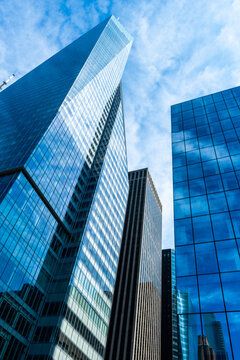 Modern skyscrapers and glass office buildings rise above an urban business district, featuring reflective facades, dramatic perspective, blue sky, and contemporary city architecture.