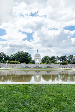 United States Capitol building viewed across a reflective pool, surrounded by green lawn and trees under a bright cloudy sky, representing government, history, architecture, and American heritage.