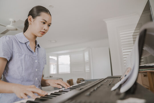piano, keyboard, girl, music, playing, home, learning. Focused student musician practices on a digital instrument in a bright modern living room with sheet music and light.