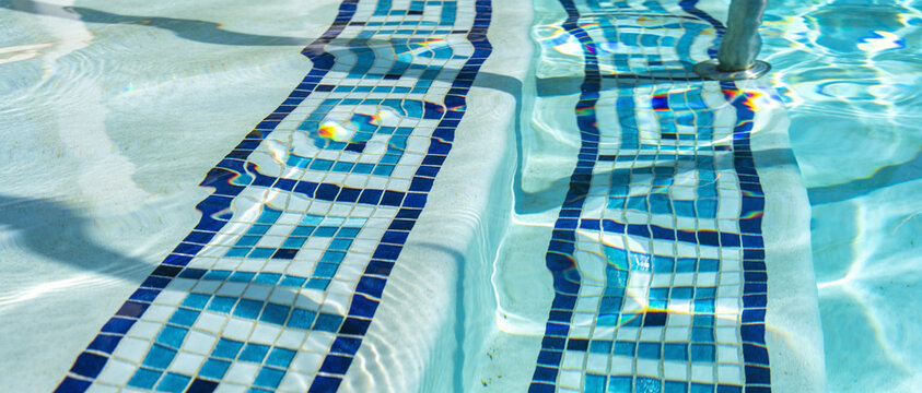 Swimming pool steps with blue mosaic tile border. Greek key pattern underwater. Sunlit turquoise water surface with ripples. Luxury resort pool details.
