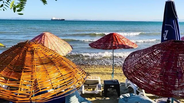 Tropical Straw Parasols and Sun Loungers on Sandy Beach with Turquoise Wavy Sea and Cargo Ship Navigating Horizon under Blue Sky on Sunny Summer Day