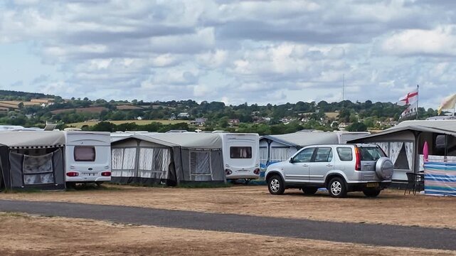 East Devon England UK. 14.08.2026. Video. A row of static caravans and motorhomes parked on a dry grassy field, with a silver SUV in the foreground, under a dramatic cloudy sky