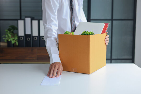 Business professional packing personal belongings into a cardboard box alongside an envelope on a desk. Concept of job resignation or termination.