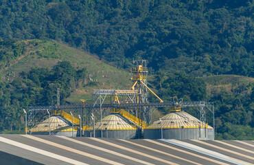 Industrial agricultural storage facility featuring metallic grain silos and conveyor systems set against a lush green hill and tropical forest backdrop © John_Doo78