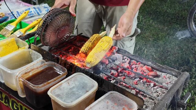 A street food vendor slowly grills fresh corn over glowing charcoal, turning each golden cob as the flames rise and smoky aromas fill the lively outdoor food stall
