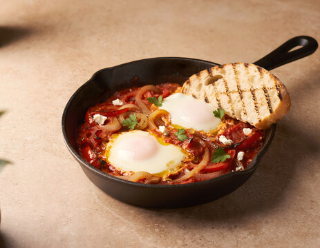 Traditional shakshuka with poached eggs in tomato sauce, rustic cast iron skillet with spicy mediterranean breakfast, middle eastern shakshuka served with grilled sourdough bread and feta cheese