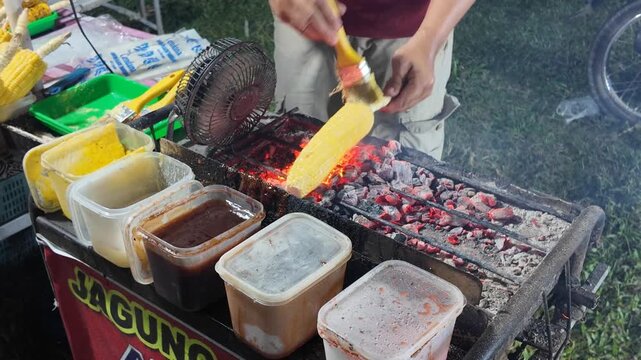 A street food vendor brushes fresh corn with flavorful sauce while grilling it over glowing charcoal, capturing the warmth, movement, and authentic atmosphere of a busy night market