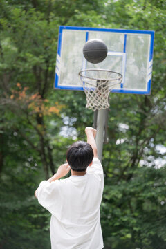 shot of a boy's back as he shoots a basketball on a lush outdoor court,without face