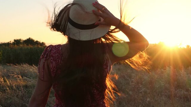 Woman from behind with long hair and a hat walking through a golden field of tall grass during a beautiful sunset, with the wind blowing through her hair and warm lens flare