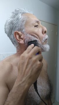 Mature man trimming and shaping his white beard and mustache with an electric clipper