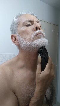 Mature man trimming and shaping his white beard and mustache with an electric clipper