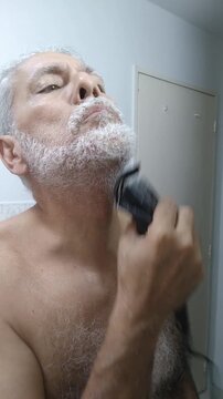 Mature man trimming and shaping his white beard and mustache with an electric clipper