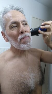Mature man trimming and shaping his white beard and mustache with an electric clipper