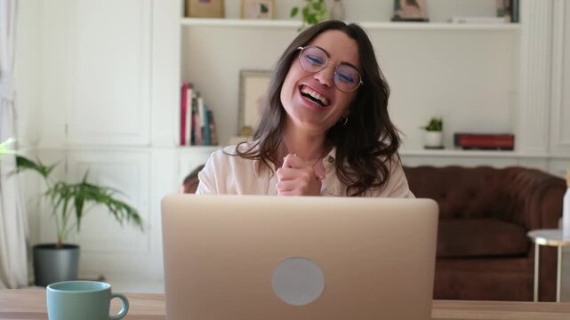 A cheerful woman in glasses reacts with pure joy and excitement while looking at her laptop. She celebrates an online success or receives good news from the comfort of her home, showing a thumbs-up.