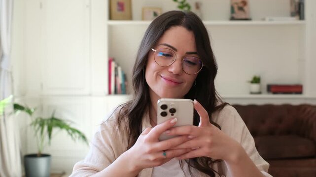 A happy young woman in glasses smiles while looking at her smartphone at home. She appears to be texting or browsing content online, reflecting with a thoughtful and pleasant expression on her face.