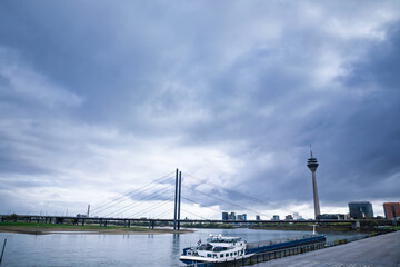 Panorama of the Dusseldorf waterfront on the Rhine, Germany, with a boat in the foreground, Rheinkniebrucke bridge crossing the river and the Rheinturm tower rising above the skyline. © Jerome