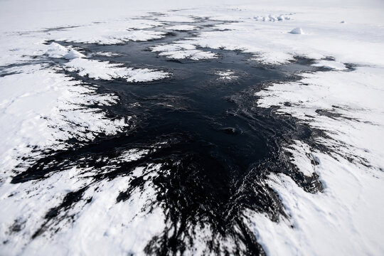 A vast white snowfield defiled by a sprawling black oil spill, aerial view.