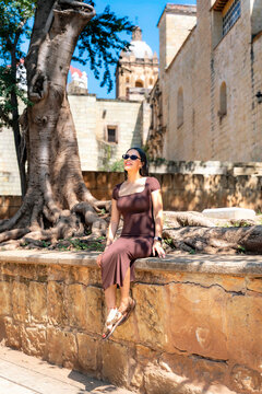 Young latina woman sitting on a stone wall, relaxing outdoors in oaxaca city with a historical building and large tree in the sunny background, experiencing travel and culture