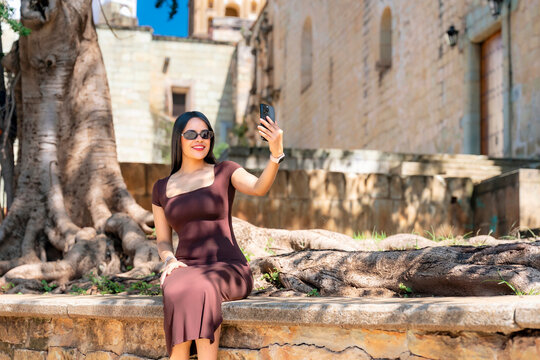 Latina woman sitting on a stone bench taking a selfie with her smartphone, smiling happily in front of an ancient building and large tree in oaxaca, mexico