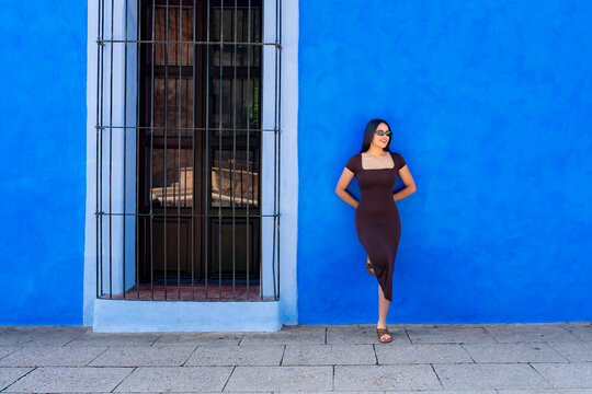 Young latina woman standing against a brightly painted blue wall in oaxaca, mexico, wearing a stylish brown dress and sunglasses, enjoying travel and urban exploration