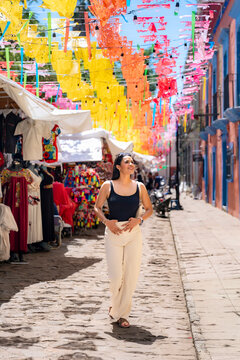 Latina woman walking along a cobblestone street adorned with colorful papel picado banners, exploring a bustling market with traditional clothing and crafts on a sunny day in oaxaca