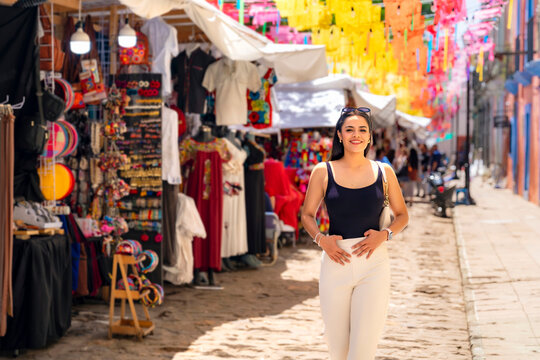 Latina woman enjoying a vibrant day, smiling while walking along a colorful street market in oaxaca, mexico, offering a glimpse into local culture and commerce