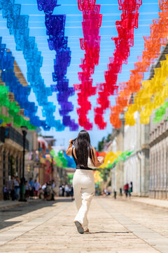 Latina woman with long dark hair walking on a cobblestone street decorated with traditional papel picado banners, exploring the vibrant culture of oaxaca, mexico