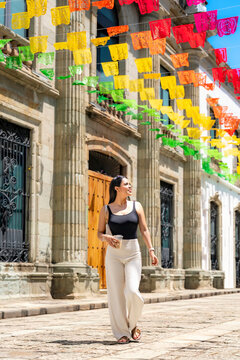Young latina woman strolling down a cobblestone street, looking up at vibrant traditional papel picado banners decorating the colonial architecture of oaxaca, mexico