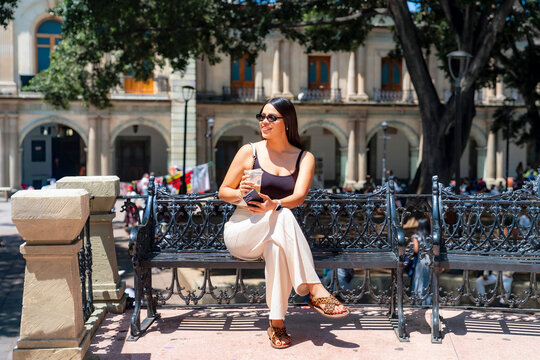 Latina woman sitting on a decorative bench in oaxaca's zocalo, smiling and sipping a cool drink while enjoying sunny, relaxed sightseeing in a historic city square
