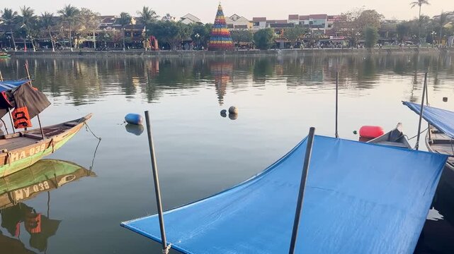 Rising camera shot moving from wooden boats in foreground to calm Thu Bon River and historic waterfront with colorful lantern tower in Hoi An Vietnam