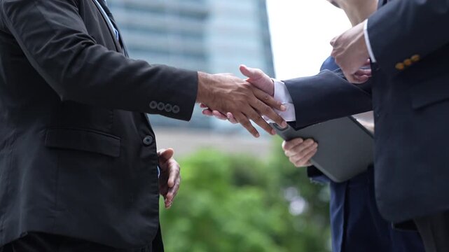 Two businessmen in dark suits shake hands in close up to confirm a business agreement under fresh trees in the public realm of a smart city district, white shirt cuffs show at the wrists