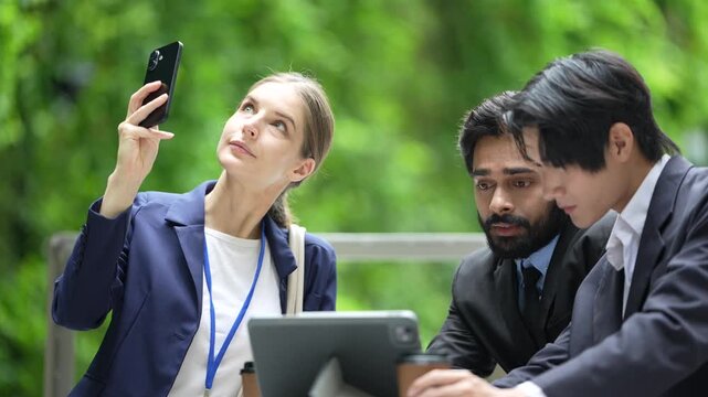 Three business colleagues compare a smartphone and a tablet at an outdoor meeting table beside the polished facade of a new mixed use city development, green hedges enclose the seating area