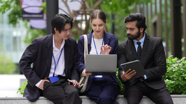 Three business colleagues share a laptop and a tablet during an outdoor team session along a sunlit walkway between contemporary corporate towers, trimmed greenery frames the seating