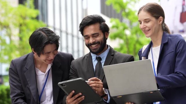 A multiethnic business team laughs at a result shown on a laptop during an outdoor stand up under fresh trees in the public realm of a smart city district, soft daylight fills the courtyard