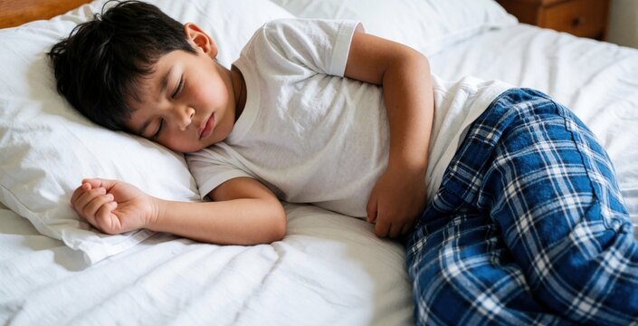 Young boy with stomach ache lies in bed. Child slepic uncomfortably, holding his belly. He looks unwell, feeling pain and discomfort at home.