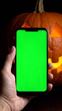 Person Holding a Smartphone with a Green Screen Chroma Key Display in Front of a Glowing Jack O Lantern Halloween Pumpkin
