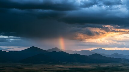 Obraz premium A small rainbow arc appears above rugged mountain silhouettes during a stormy, golden hour sunset
