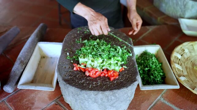 Experienced chef finely chopping fresh herbs with a knife on a traditional stone metate, preparing ingredients for a delicious mexican salsa with tomatoes and other fresh vegetables