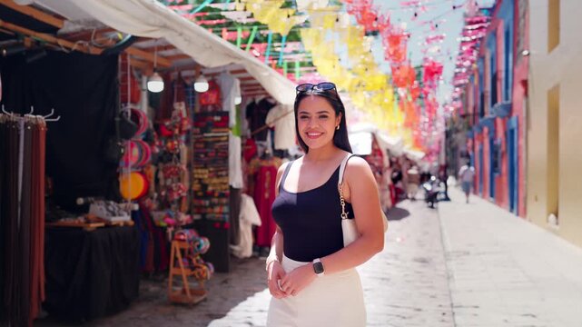 Smiling Latina woman enjoying a sunny day while walking along a vibrant street market in Oaxaca, Mexico, surrounded by colorful decorations and local crafts during her vacation