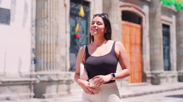 Stylish Hispanic woman with a coffee walking along a sunny, historic street in Oaxaca, Mexico, looking around and smiling happily at the camera while exploring the beautiful colonial city