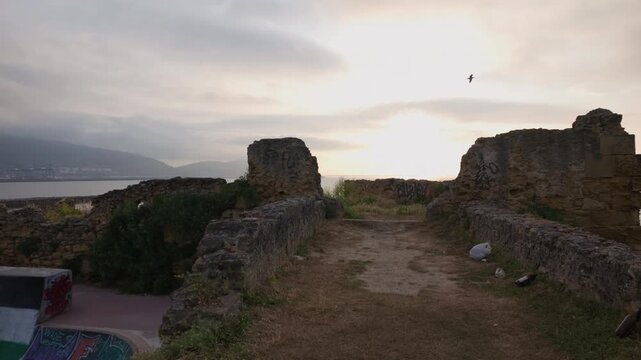 Old stone ruins on a coastal hill overlooking the ocean at sunset, featuring a graffiti covered skate ramp and mountains