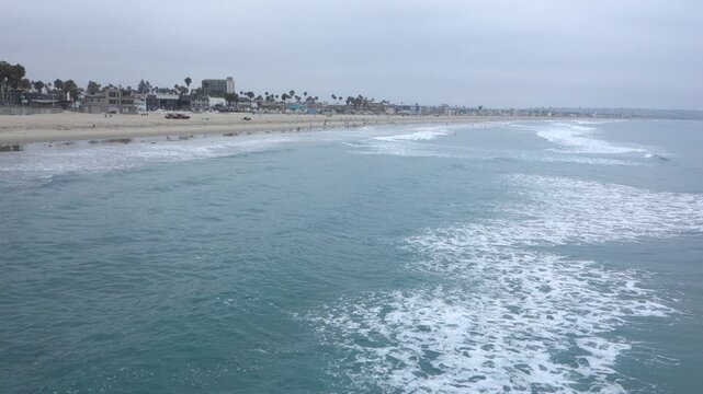 Bird-eye view of foaming turquoise ocean waves approaching a wide sandy Pacific beach shoreline with vacation-friendly rentals and hotels stretching along the coast, San Diego, California