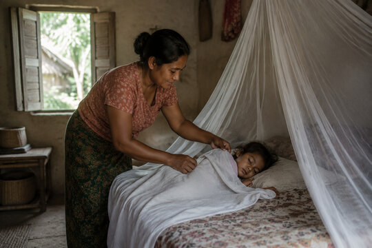 Mother tucking child under white mosquito net in simple rural bedroom, malaria prevention and maternal care concept.