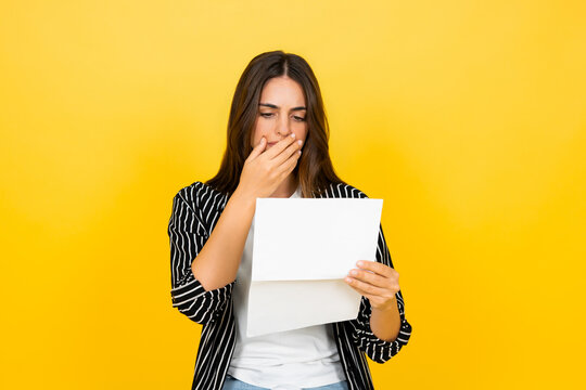 Woman standing on a yellow background, holding an open letter and covering her mouth in shock and disbelief, expressing emotions of surprise, sadness, and bad news