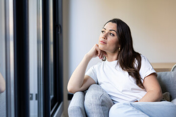Fototapeta premium Young woman sitting comfortably on a sofa at home, resting her chin on her hand, looking thoughtfully and reflectively out a large window, lost in deep contemplation and daydreaming