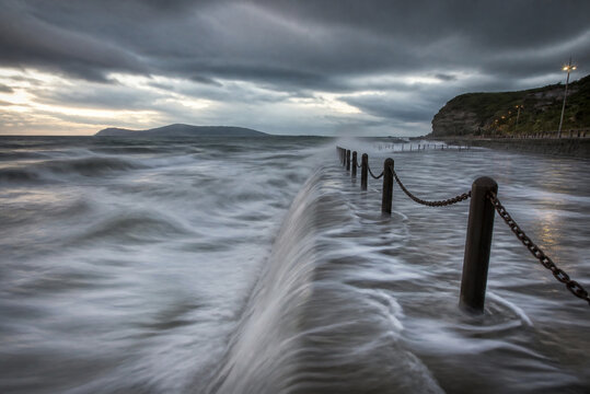 Ocean waves overflowing a concrete seawall, coastal landscape with dramatic weather and long exposure.