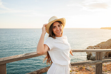 Fototapeta premium Young woman smiling wearing a straw hat and white outfit, standing by a wooden fence overlooking the vast ocean during a beautiful golden hour sunset, enjoying summer travel and vacation
