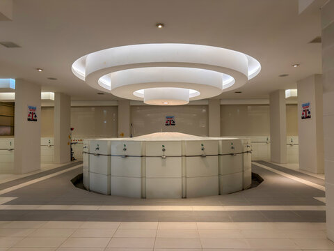 Putrajaya, Malaysia. August 24, 2026. A modern interior featuring a circular white ablution station under a multi-tiered ceiling light fixture, surrounded by clean white walls, tiled flooring, and nea