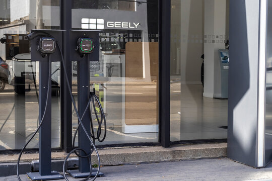 Two smart electric vehicle charging units, equipped with cables and plugs, are mounted on pedestals against the glass facade of a Geely dealership in the city of S&atilde;o Paulo. 