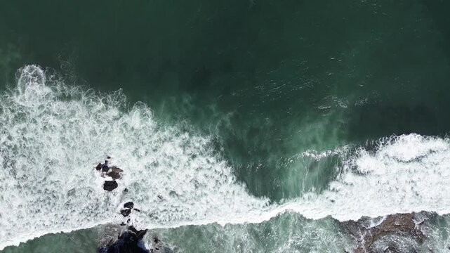 Aerial view shows waves crashing against rocks. Deep green water churns with white foam. Rocks jut from the sea, battered by surf. Nature's power is raw and relentless